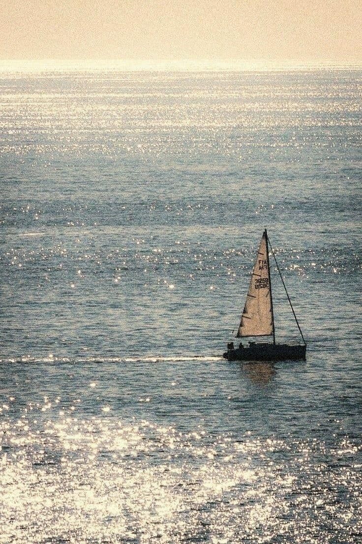 a small sailboat floating in the middle of the ocean with sun reflecting off the water