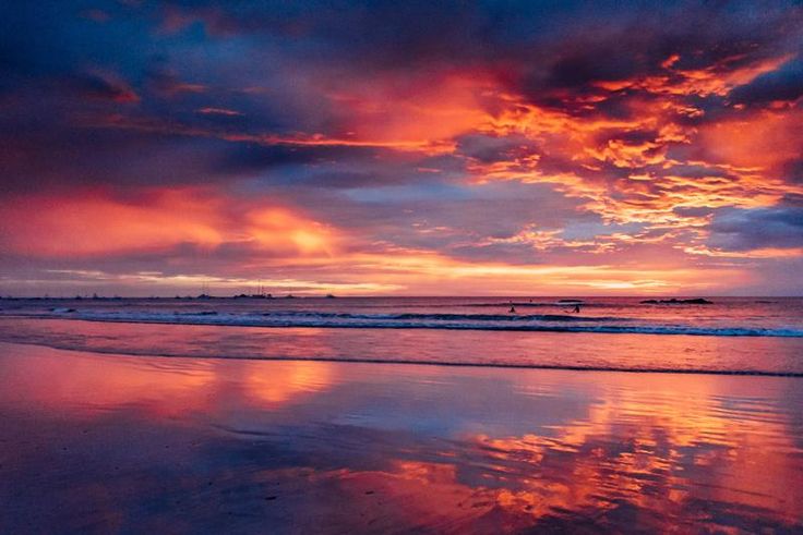 the sky is reflected in the wet sand on the beach at sunset, with people walking along the shore