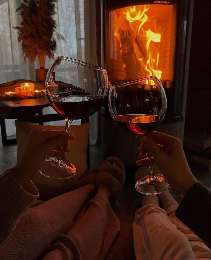 two people toasting with red wine in front of an open fire place at night