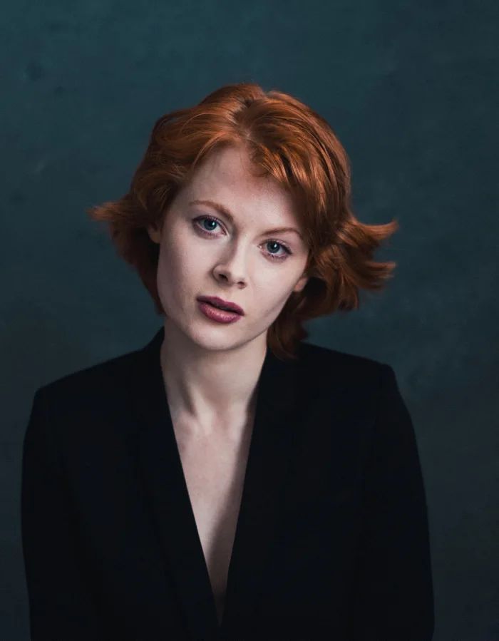 a woman with red hair wearing a black suit and looking at the camera while standing in front of a dark background