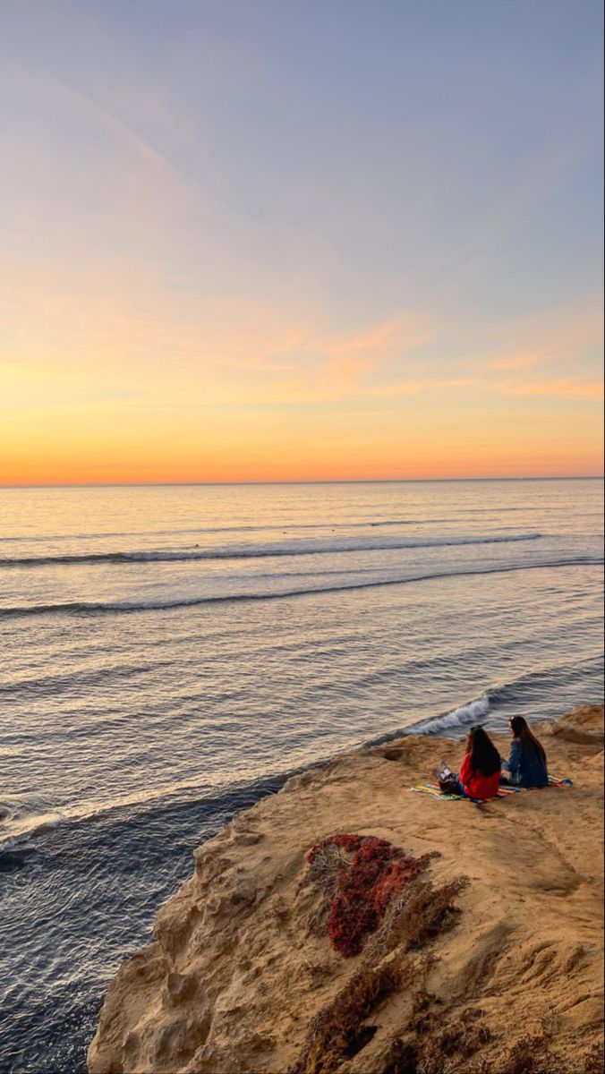 two people sitting on the edge of a cliff overlooking the ocean at sunset or sunrise