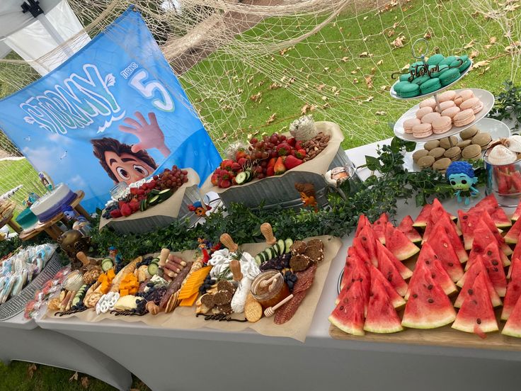 watermelon slices and other foods are on display at an outdoor party with a sign in the background