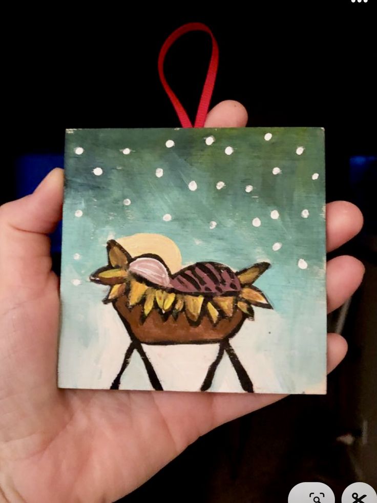 a hand holding up a small square ornament with a painting of a baby jesus in the manger