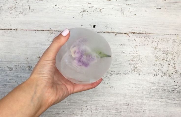 a woman's hand holding a glass bowl with flowers in it on a white wooden surface