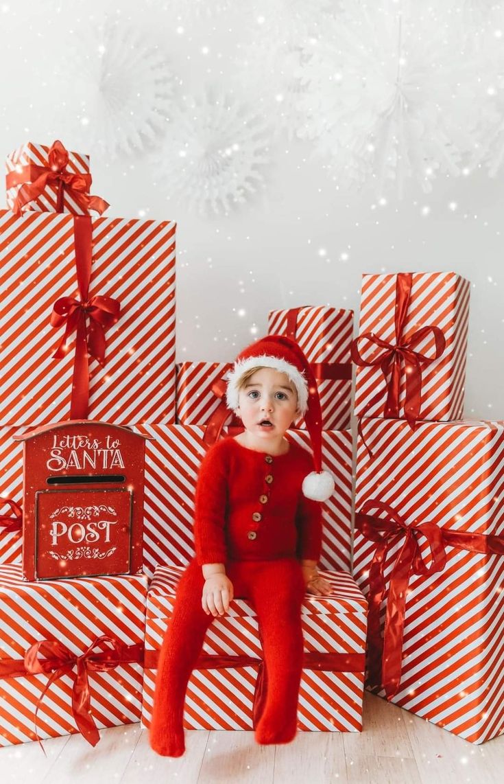 a baby sitting in front of christmas presents wearing a santa claus hat and red pajamas