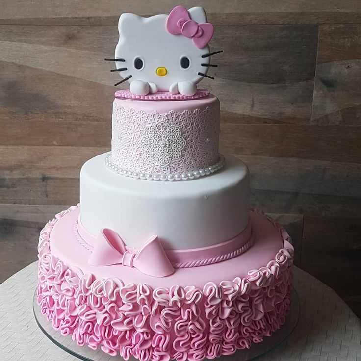 a hello kitty cake with pink ruffles and white frosting on top is displayed in front of a wooden wall