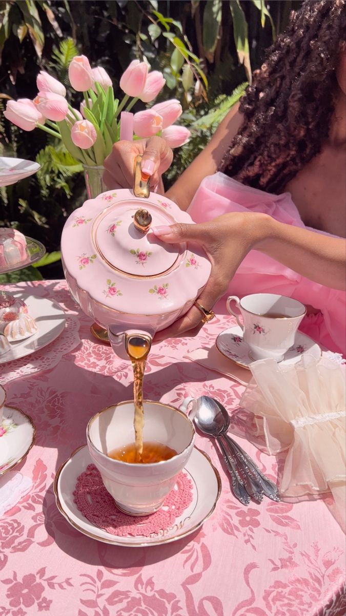 a woman pouring tea into a cup on top of a pink table cloth