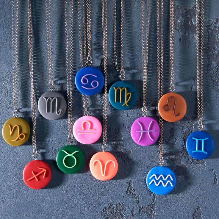 twelve zodiac signs are displayed on different colored discs hanging from chain necklaces against a gray wall