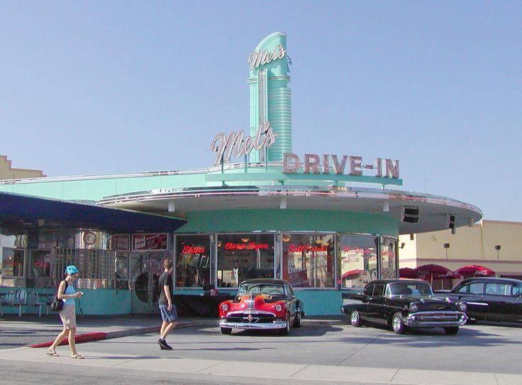 two people walking in front of a drive - in with cars parked on the street