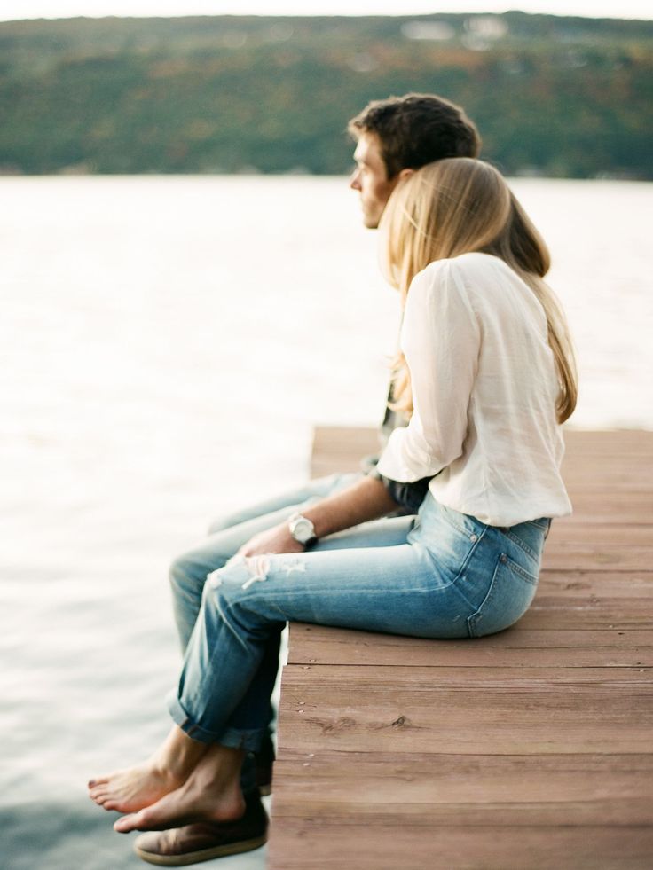 a man and woman sitting on a dock next to the water looking at each other