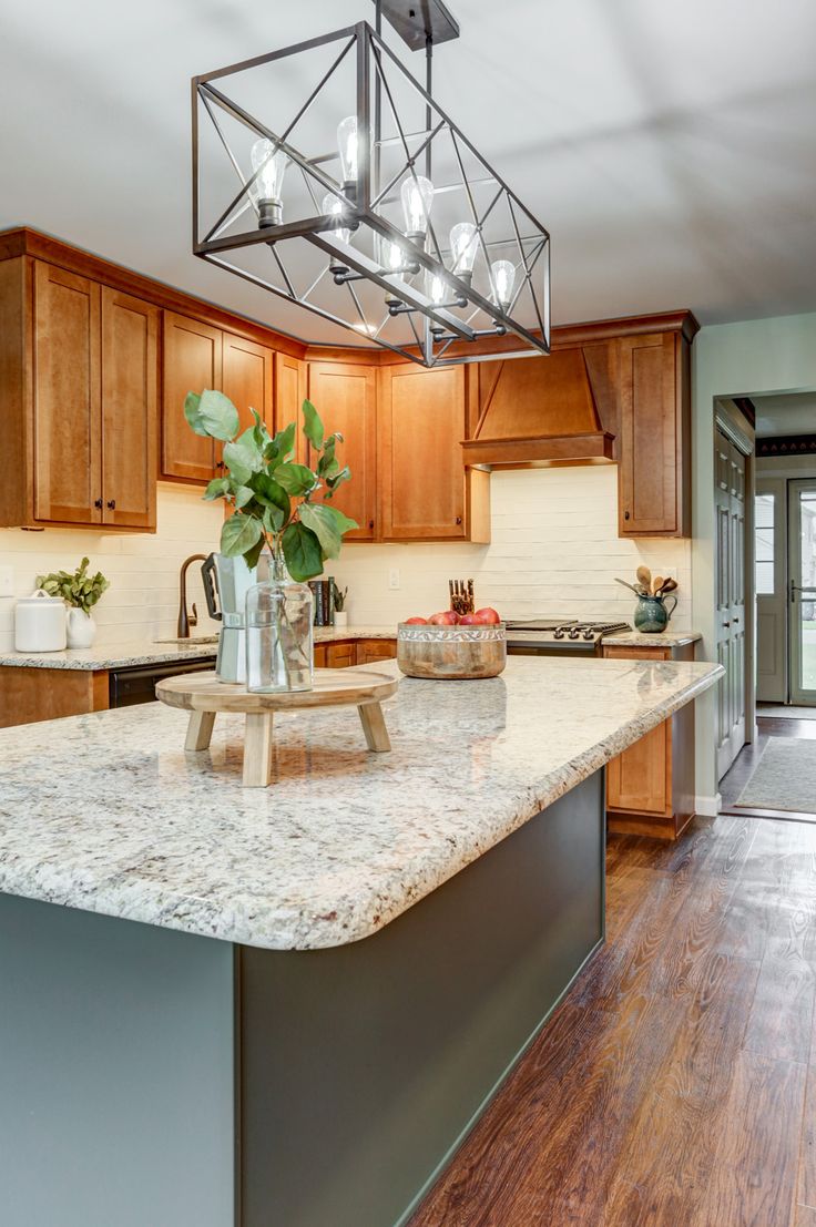 The contrasting warm and cool elements in this kitchen draw you in and makes you want to stay awhile. With white cabinets being so popular, these clients decided to go a different direction with brown maple wood wall cabinets. Adding a green island, light countertops, a white backsplash, and dark accents creates multiple textures, giving this kitchen dimension. The wall cabinets extend all the way to the large window, drawing in natural light, and the island offers bar seating. Green Kitchen Island, Honey Oak Cabinets, Maple Kitchen Cabinets, Brown Kitchen Cabinets, Light Wood Cabinets, Green Island, Oak Kitchen Cabinets, Brown Cabinets, Kitchen Wall Colors