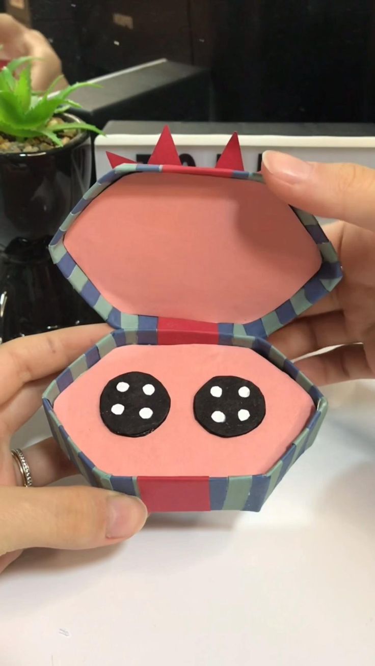 a person is holding a pink box with black eyes and horns on it, while another hand holds the lid