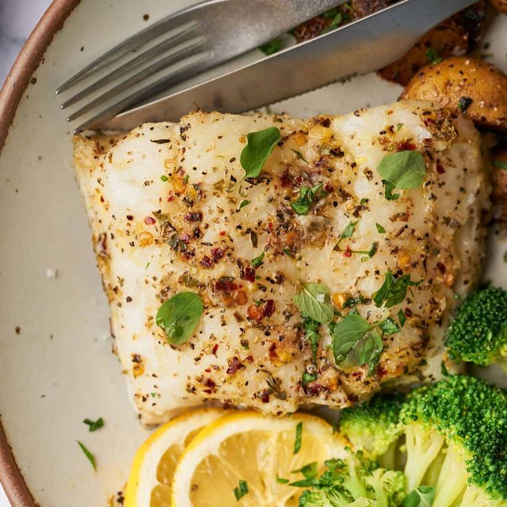 a white plate topped with fish, broccoli and lemon wedges next to a fork