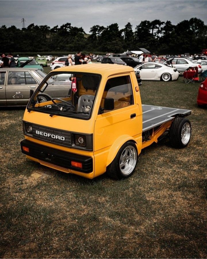 a small yellow truck parked on top of a grass covered field next to other cars