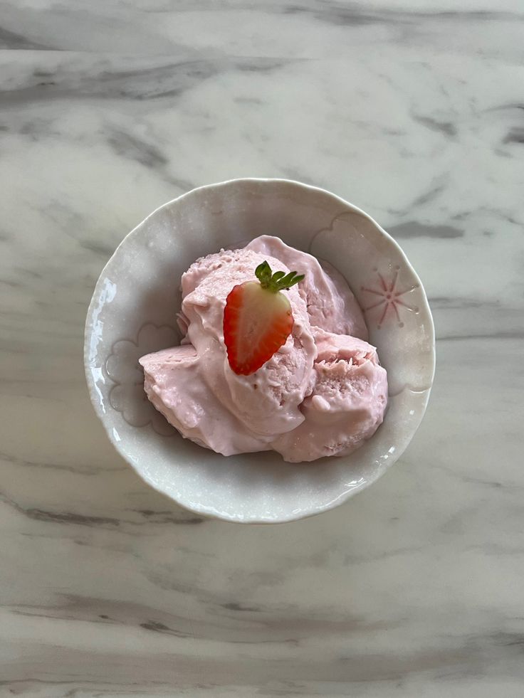 a white bowl filled with pink ice cream and a strawberry on top, sitting on a marble surface