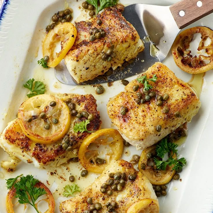 chicken with lemons, capers and olives on a white platter next to a knife