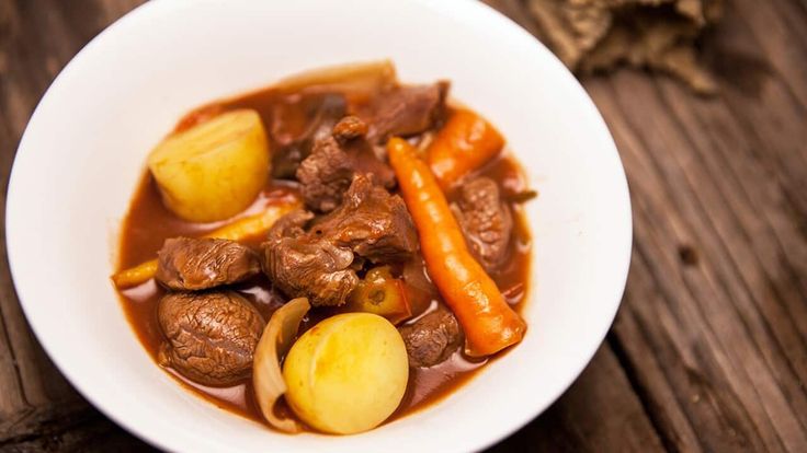 a white bowl filled with stew and carrots on top of a wooden table next to a piece of bread