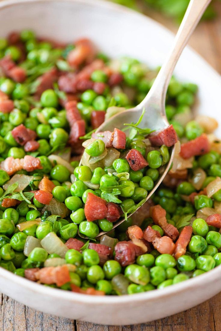 a bowl filled with peas and bacon on top of a wooden table next to a spoon