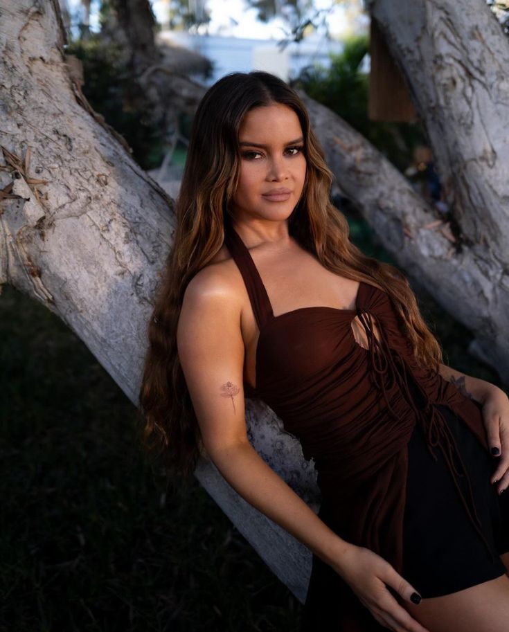 a beautiful young woman in a brown dress leaning against a tree