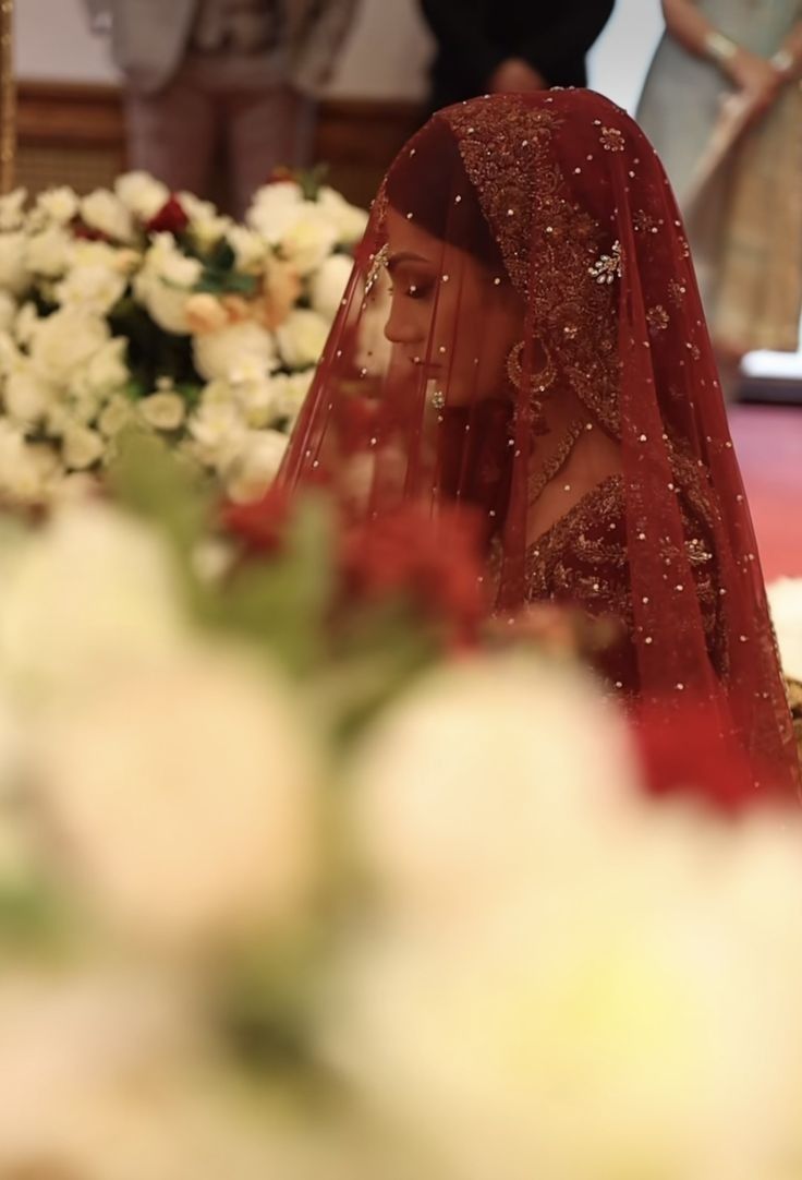 a woman in a red veil sitting at a table with flowers and people behind her