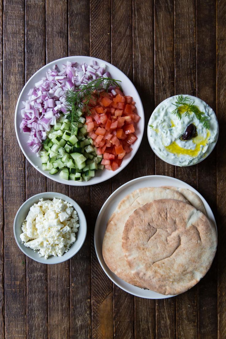 an assortment of different foods are arranged in bowls on a wooden table, including pita bread and cucumbers