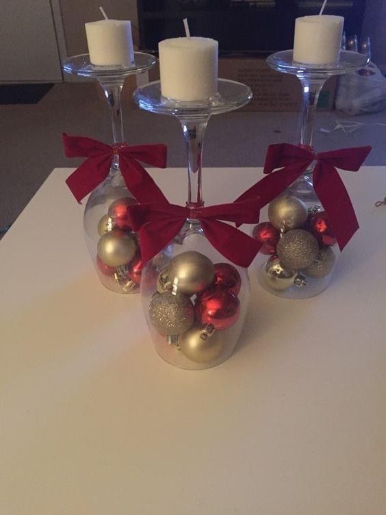 three wine glasses with christmas ornaments in them on a white tablecloth and two candles are next to each other