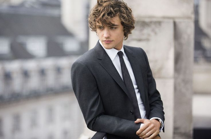 a young man in a suit and tie leaning against a wall with his hand on his pocket