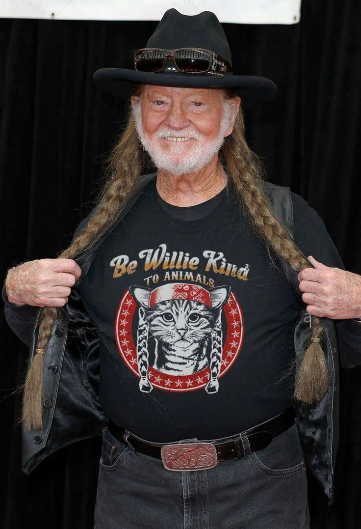 an older man with long hair wearing a black shirt and cowboy hat, holding up his t - shirt