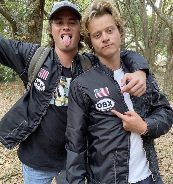 two young men standing next to each other in the woods with trees behind them and one man sticking out his tongue