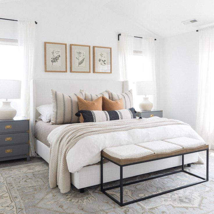 a white bed with pillows and blankets on top of it in a bedroom next to two windows