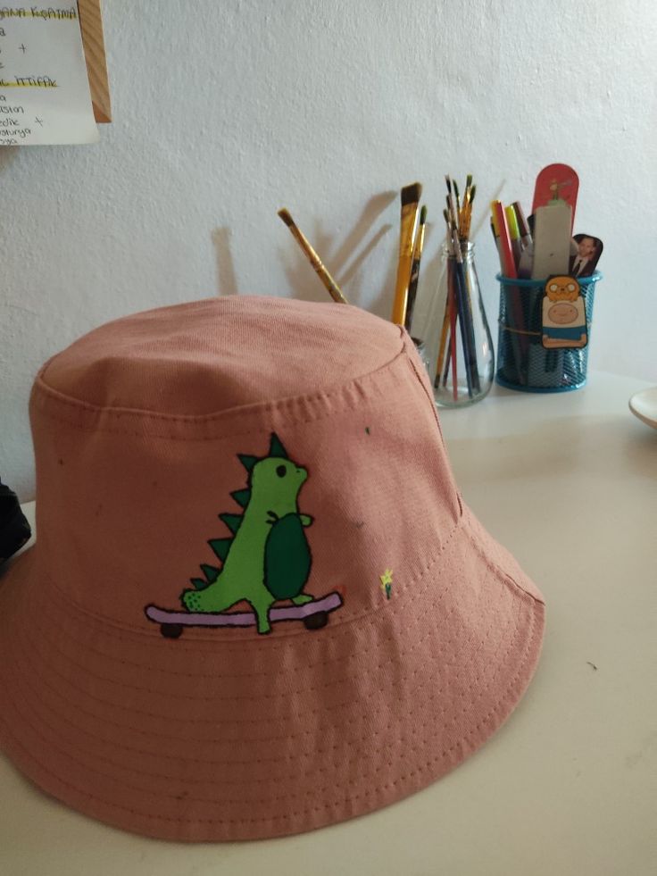 a pink bucket hat with a green dinosaur on the front, and writing utensils behind it