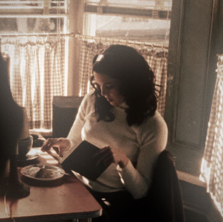 a woman sitting at a table in front of a window reading a book and talking on the phone