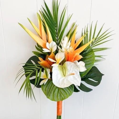 an arrangement of tropical flowers and palm leaves in a vase on a wooden table against a white wall