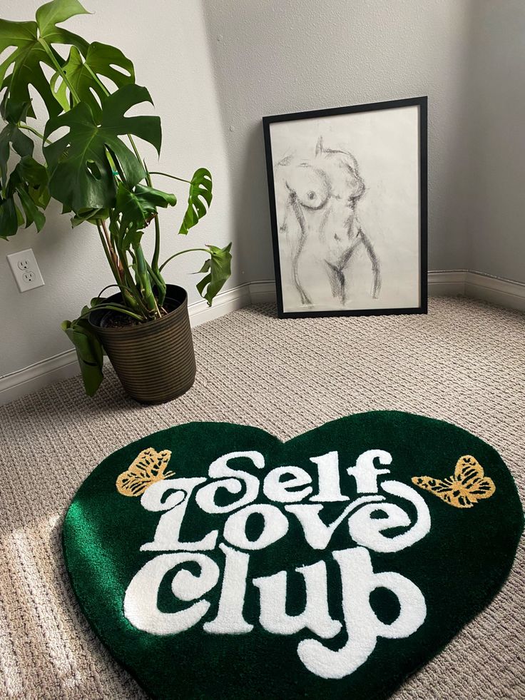 a green heart shaped rug with the words self love club on it next to a potted plant