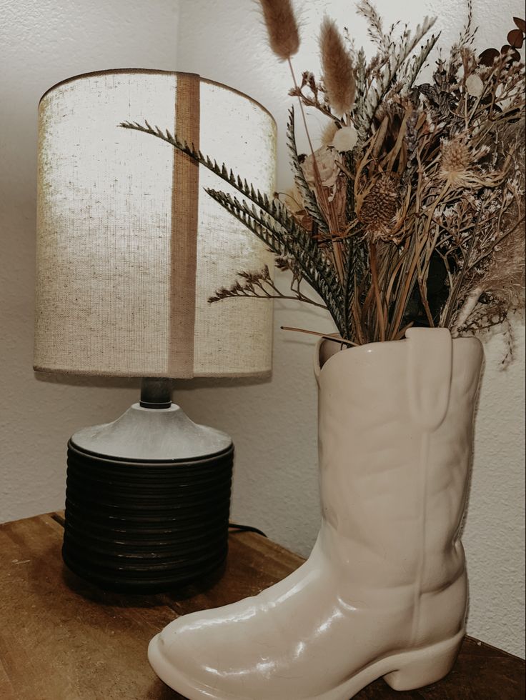 a white cowboy boot next to a lamp on a wooden table with dried plants in it