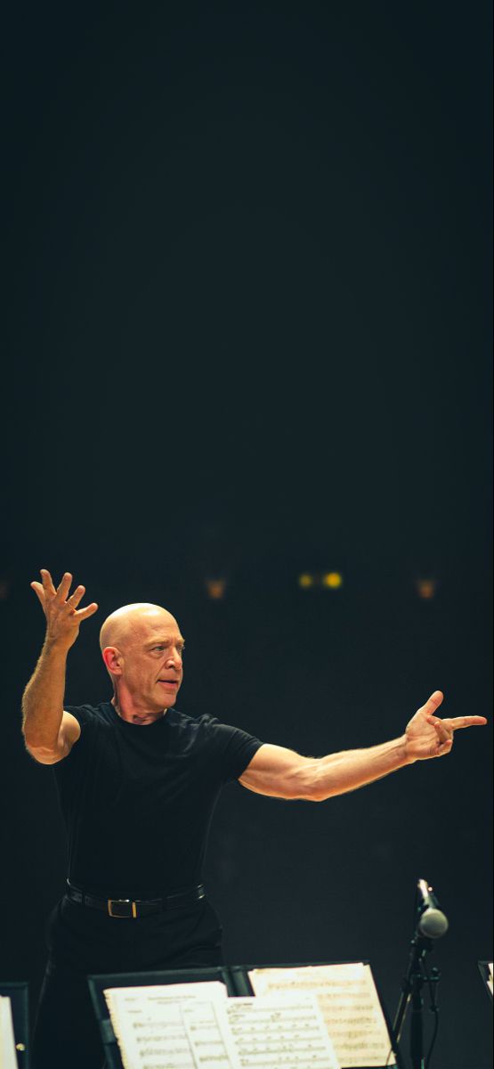 an older man with his arms outstretched in front of music sheets and microphones on stage