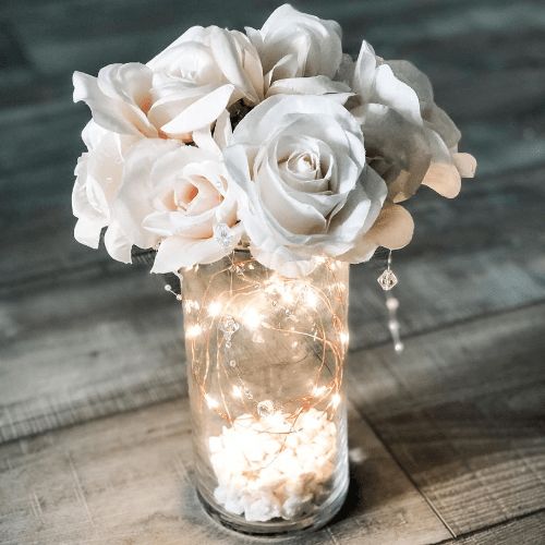 a glass vase filled with white flowers on top of a wooden table covered in fairy lights