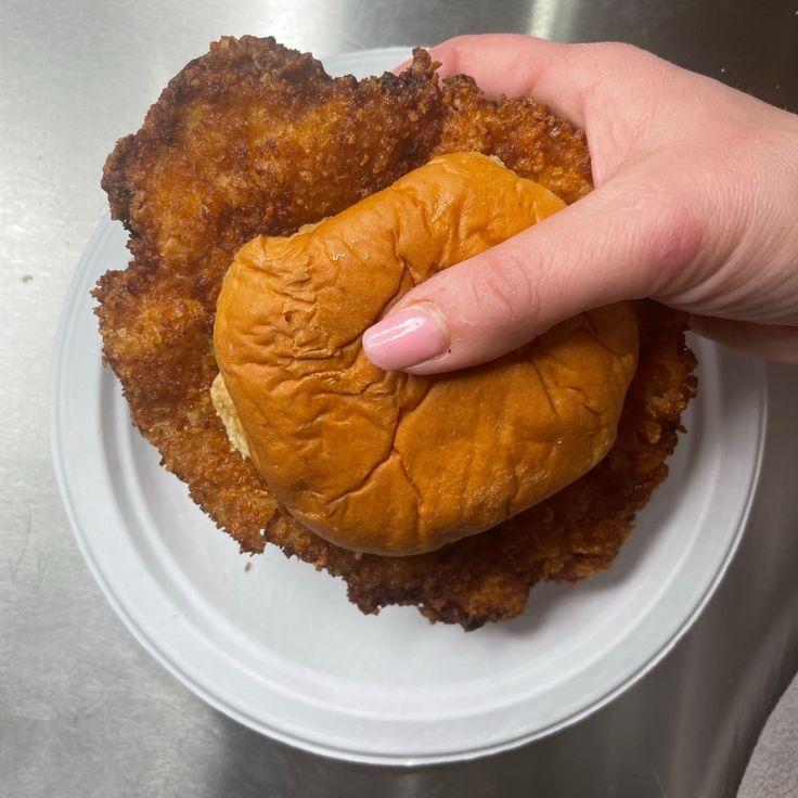 a person is holding a piece of fried chicken on a bun in their left hand