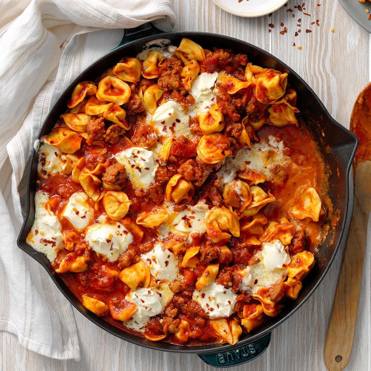 a skillet filled with pasta and sauce on top of a wooden table next to a spoon