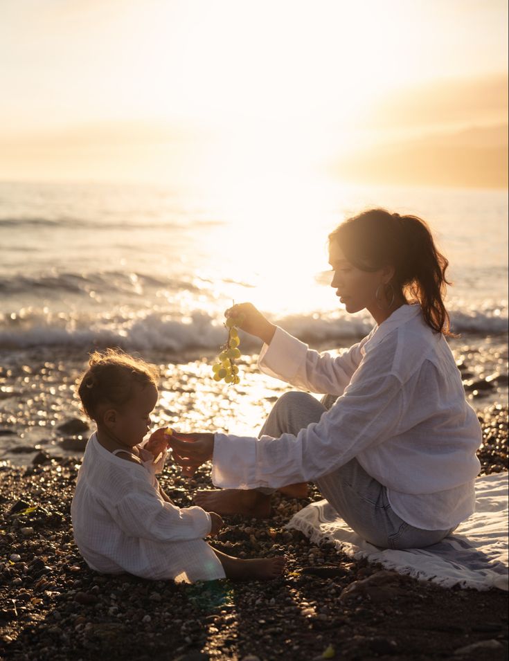 a woman and child sitting on the beach at sunset, one is holding a flower