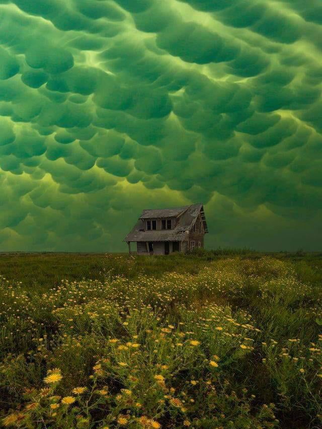 a house sitting in the middle of a field under a green sky filled with clouds