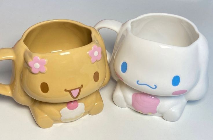 two ceramic mugs sitting next to each other on a white counter top, one has a cat and the other has a flower in it's mouth