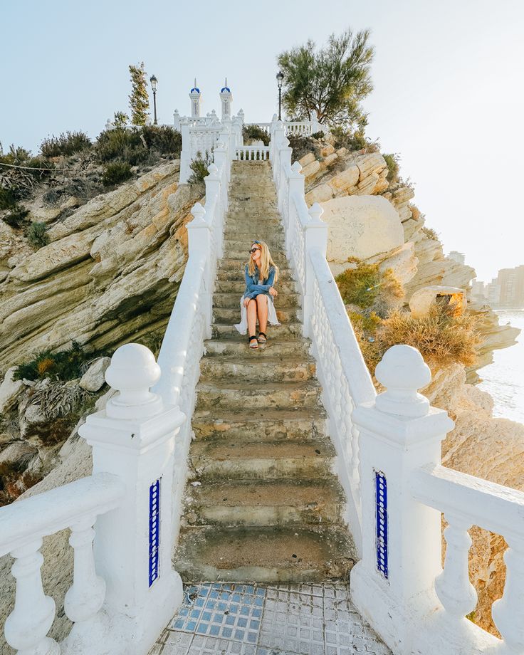 a woman sitting on top of a set of stairs next to the ocean with her legs crossed