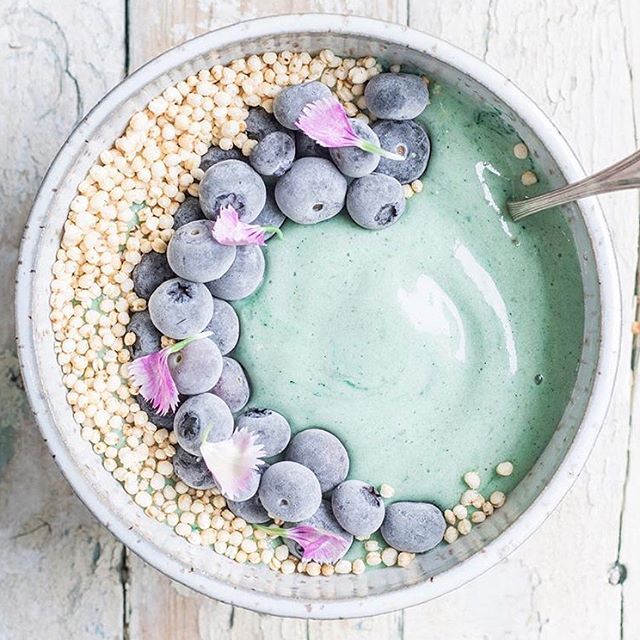 a bowl filled with blueberries and corn on top of a white wooden table next to a spoon