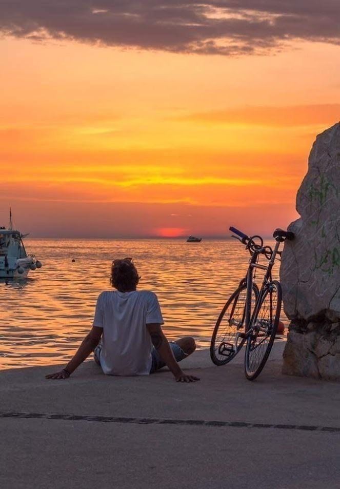 a person sitting on the ground next to a bike near water with boats in the background