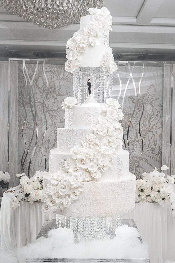 a large white wedding cake with flowers on the side and chandelier hanging from the ceiling