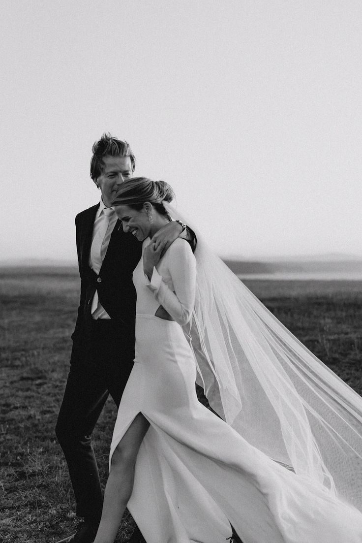 a bride and groom walking in the grass together with their veil flying over their heads