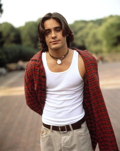 a young man with long hair wearing a white tank top and brown pants standing in front of a brick road