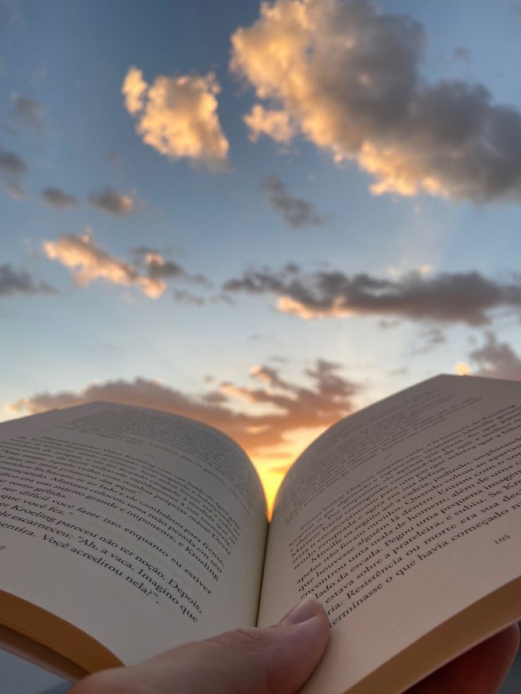 a person is holding an open book in their hand as the sun sets behind them