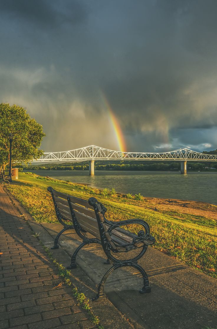 a bench sitting on the side of a road next to a river under a rainbow